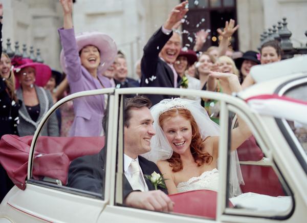 Bride and groom in convertible car, wedding party waving in background